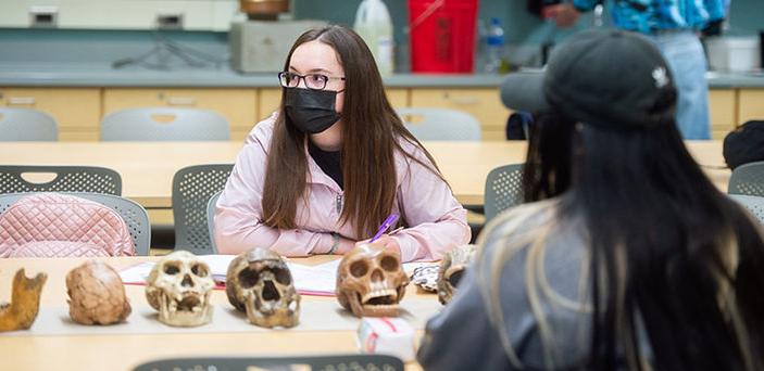 2 students sit at a table in front of a row of skulls listening to the professor and taking notes